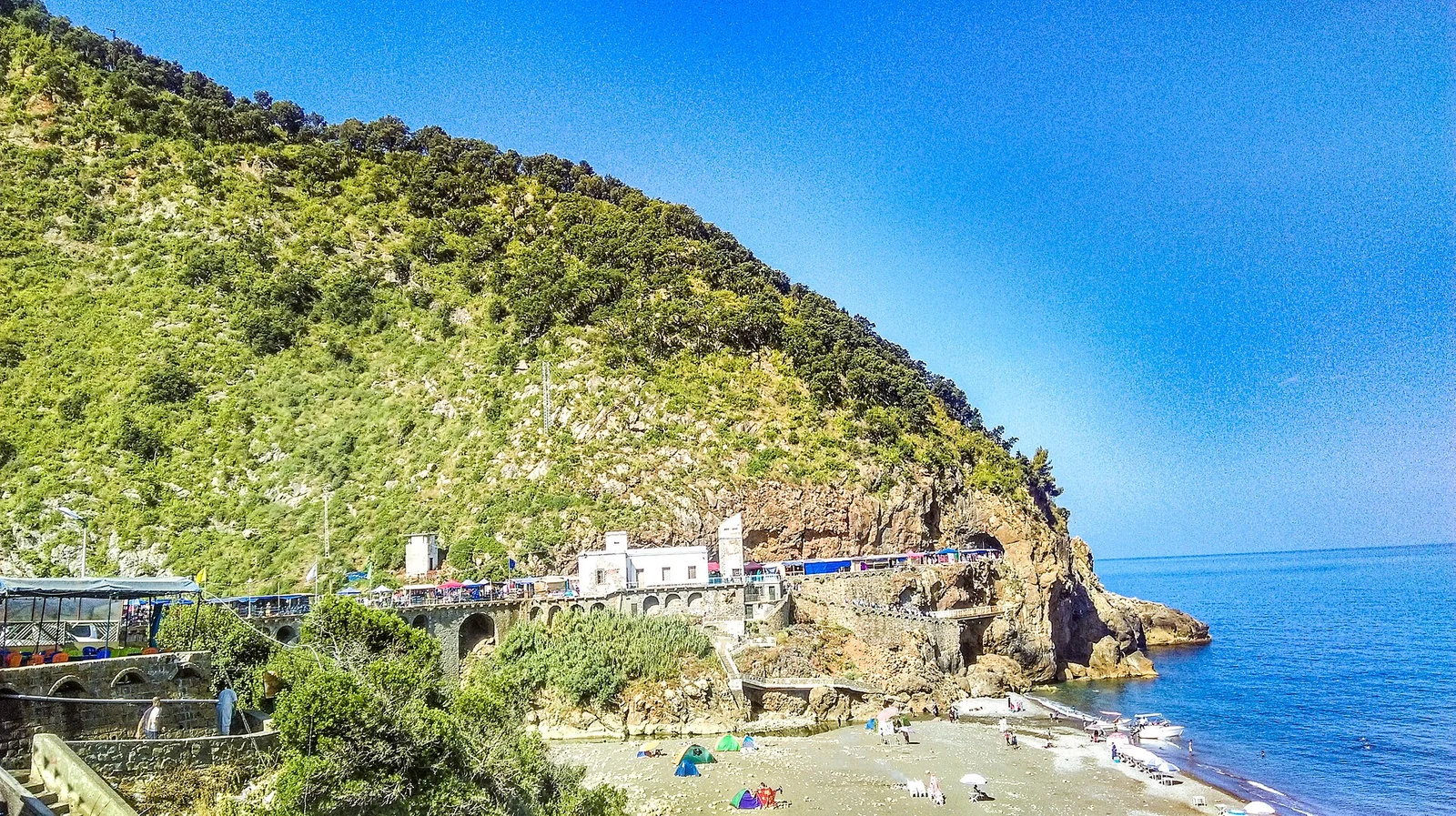 Un promontorio verde sobre una carretera costera, una pequeña playa y el mar azul.